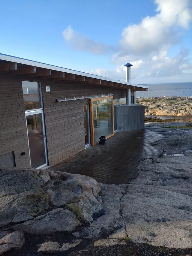 Kystnær hytte med trekledning, store vindusflater og terrasse på svaberg ved sjøen.