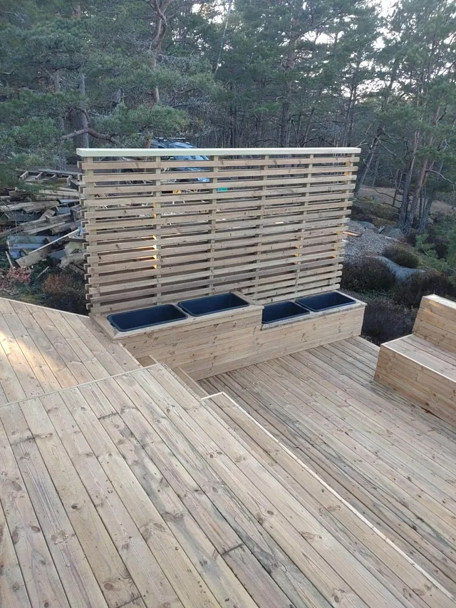 Treterrasse med levegg, plantekasser og sittebenk omgitt av furuskog og svaberg.