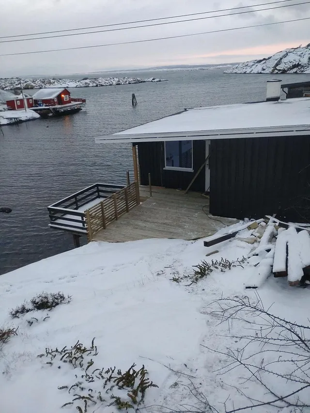 Kysthytte med terrasse ved sjøen, omgitt av snø, svaberg og små naust i bakgrunnen.