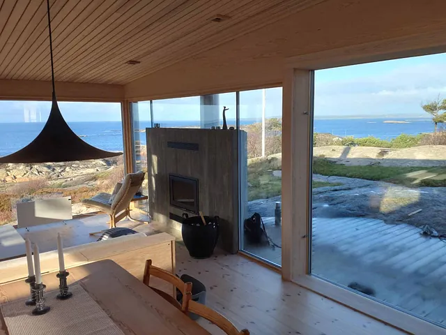 Lys stue med peis, store vindusflater og panoramautsikt mot sjø og kystlandskap.