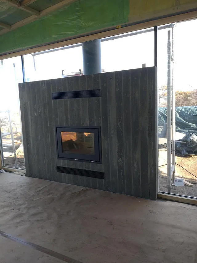 Moderne peis integrert i steinkledd vegg mellom store gulv-til-tak-vinduer under bygging.