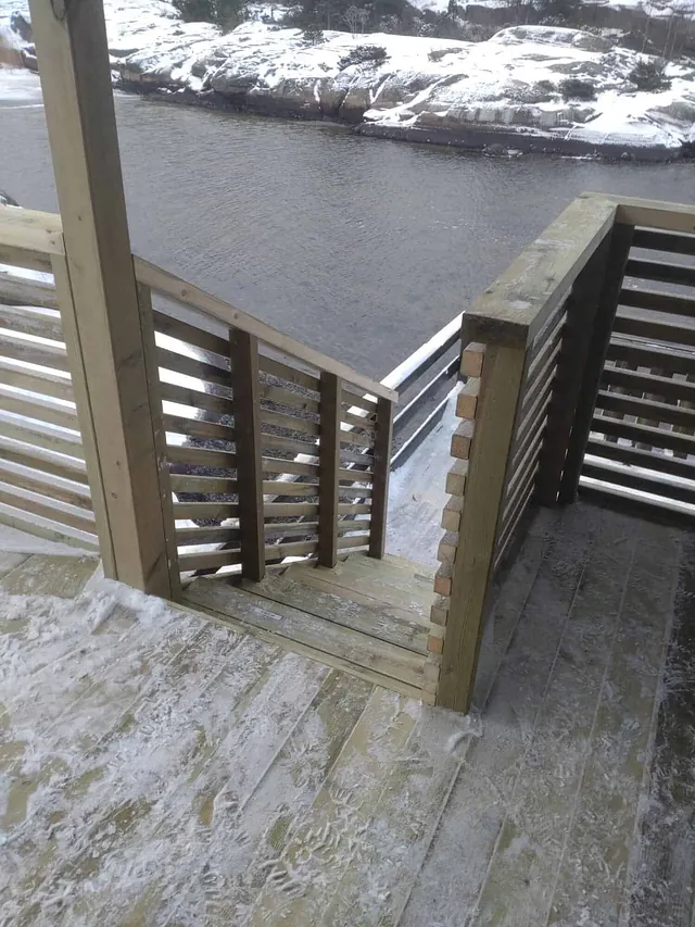 Trapp og terrasse i tre med rekkverk, plassert ved sjøkant omgitt av snødekt kystlandskap.
