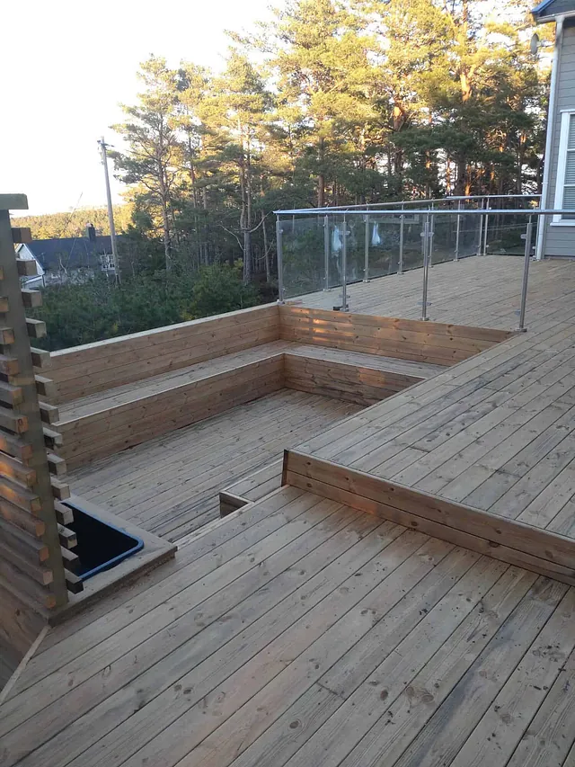 Stor treterrasse i flere nivåer med sittebenker og glassrekkverk ved enebolig i skogkant.